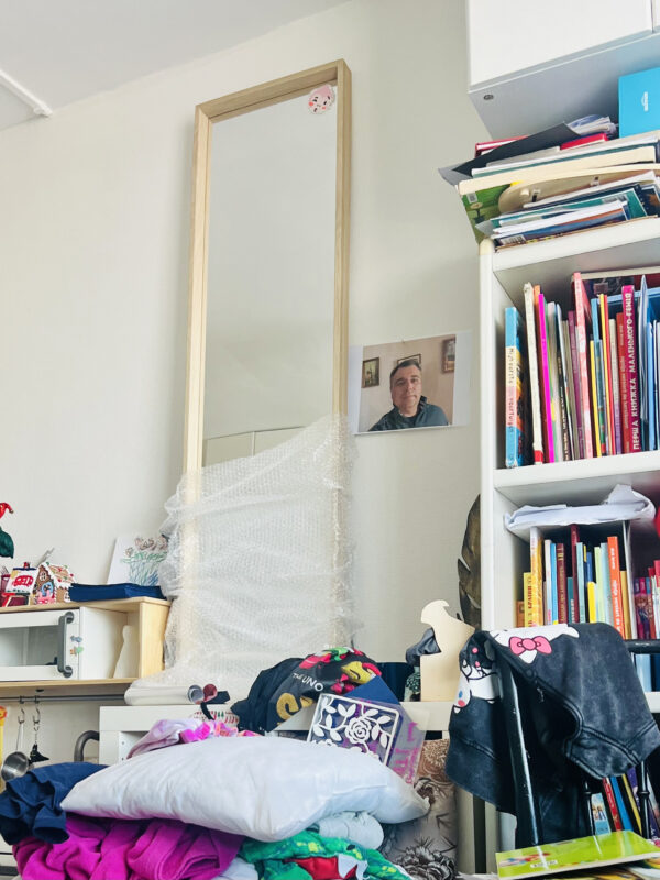 A children's bedroom. A mirror is half covered in bubble wrap with a pile of clothes and cushions in front. On the left is a bookcase filled with colourful books. A printed paper of an older man is stuck to the wall in between.