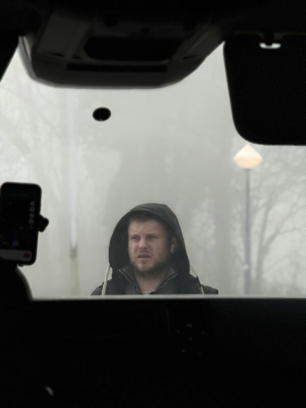 A man stands outside in the mist, hood up with a concerned look on his face, captured from inside a dark car.