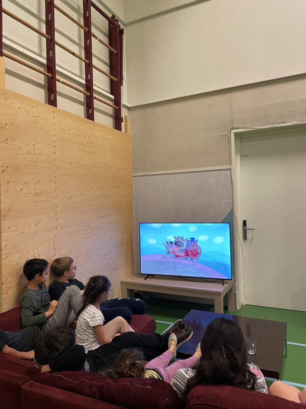A group of children sit on a red sofa, watching a cartoon on a television.