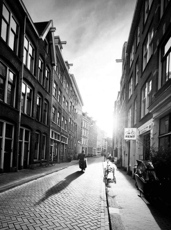 A black and white photo of a street in the centre of Amsterdam with a solitary cyclist in the road. The sun faces the camera, casting a long shadow of the person.