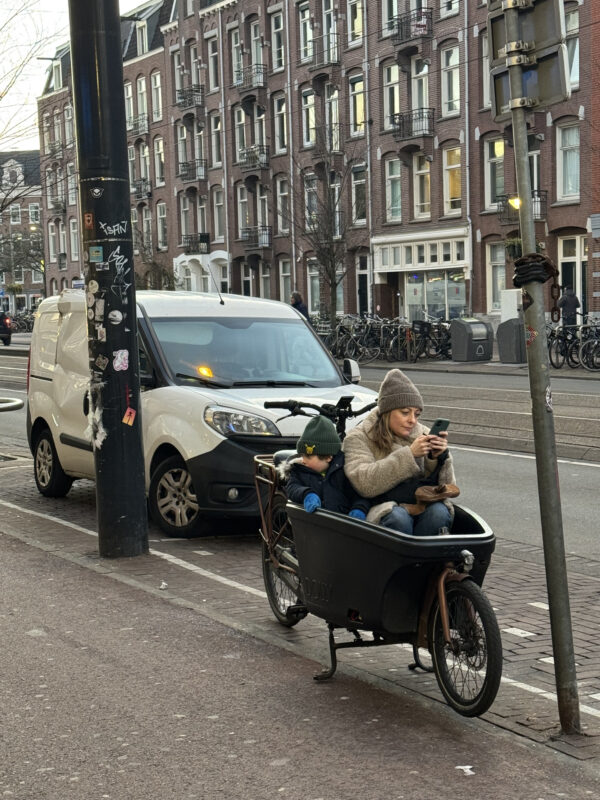 On a bike lane in Amsterdam, a mother sits in a bakfiets with her child looking at her phone.