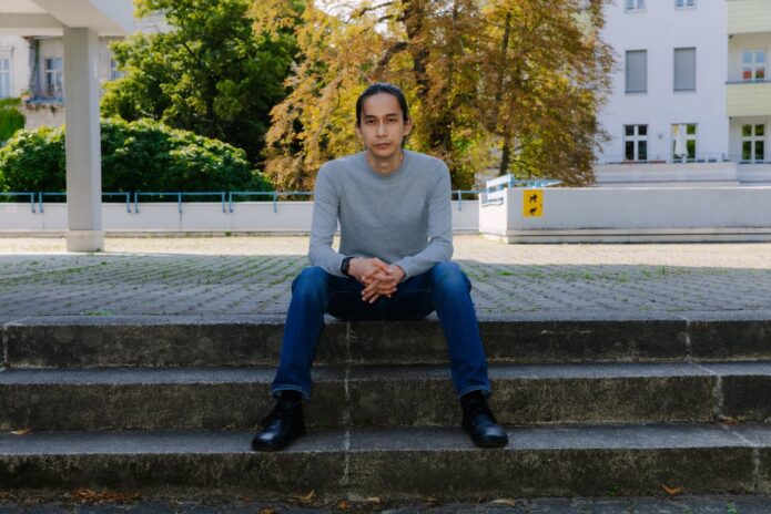Jay Afrisando, a medium-skin-toned male with bunned hair, sits on outdoor neighborhood concrete steps in a serious pose, with his fingers clasped and arms on his thighs. He wears dark blue jeans, a light gray sweatshirt, and black shoes. Photo: © DAAD Artists-in-Berlin Program / Diana Pfammatter.