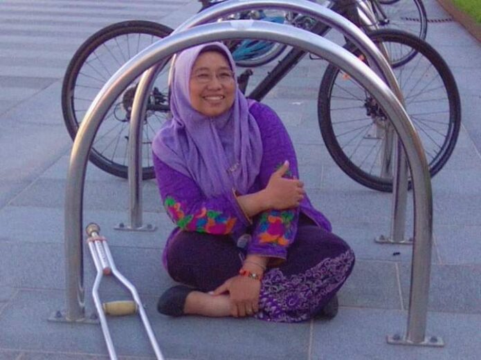Rini Rindawati, a medium-skin-toned woman, sits cross-legged on an outdoor concrete floor with a bike rack as her visual frame. She wears a purple hijab and tunic. Her crutch lies beside her.