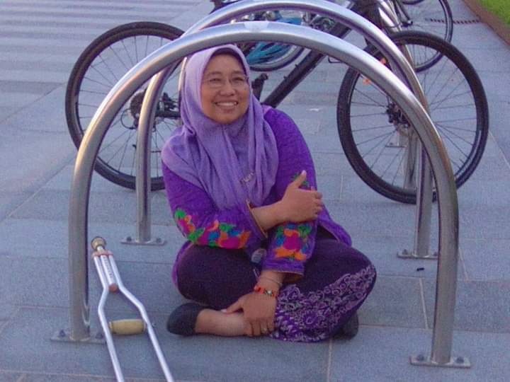 Rini Rindawati, a medium-skin-toned woman, sits cross-legged on an outdoor concrete floor with a bike rack as her visual frame. She wears a purple hijab and tunic. Her crutch lies beside her.