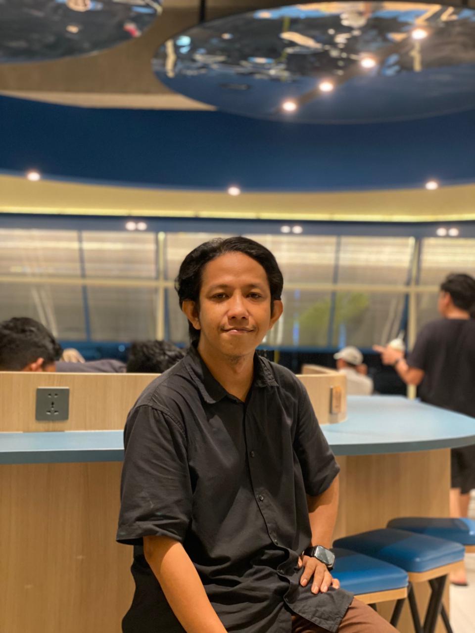 Arief Wicaksono, a medium-dark-skin-toned man with short hair, sits on a blue high stool. He wears a black shirt and a watch on his left hand. Behind him are tables and people scattered in space.