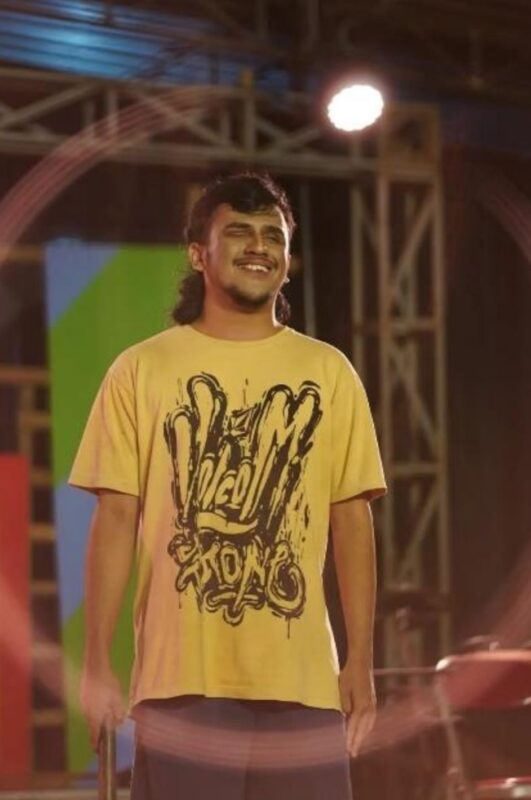 Yuda Wirajaya, a medium dark-skin-toned man with shoulder-length bunned hair, stands on stage and smiles. He wears a branded yellow t-shirt and holds a cane.
